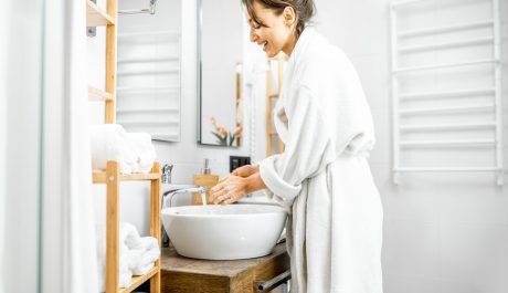 Young and cheerful woman dressed in white bathrobe having some hygienic procedures, washing hands in the bathroom at home